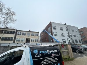 Urbana Dryer Vent Cleaning company van in the foreground with a boom lift in the background with an employee cleaning a dryer vent on a multi unit condo apartment building
